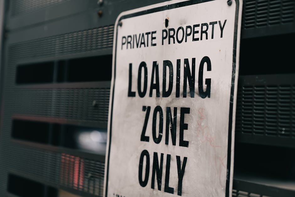 A close-up image of a metal sign mounted on a dark, perforated surface, indicating 'Private Property Loading Zone Only' in bold black letters. The sign appears weathered, with some minor rust or dirt marks, and is positioned outdoors, likely on a building's exterior near a designated loading area. The background features horizontal slats or vents, suggesting an urban environment relevant to moving and furniture transport logistics. This signage emphasizes restricted access for loading purposes, which is important for house removals and professional relocation services such as those offered by Man and Van Mayfair. The image visually supports the context of loading zones in residential or commercial properties within the Mayfair area, aligning with UK parking and loading rules.