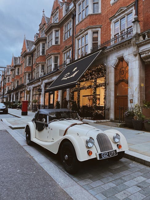 A vintage cream-colored convertible car with a black soft top, parked on a street in Mayfair, London, facing the viewer's left. The car has a rounded grille, large circular headlights, and a UK license plate reading G12 UVB. It is situated next to a building with a brick façade and white stone architectural details, including arched windows and decorative cornices. A black outdoor awning with white lettering covers a storefront entrance, which is visible behind the car. The street is paved with cobblestones and modern pavement, with a sidewalk running alongside the buildings. To the right, there are potted plants and a door with the number 13. This scene, associated with house removals and relocation services, illustrates the urban environment where furniture transport and packing processes may take place, as managed by Man and Van Mayfair.