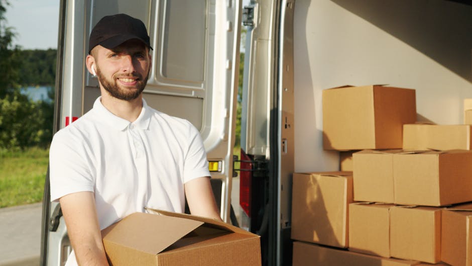 A young man with a beard, wearing a black cap and a white polo shirt, standing with his arms crossed inside the open rear of a moving van. Behind him, there are several cardboard boxes and plastic-wra