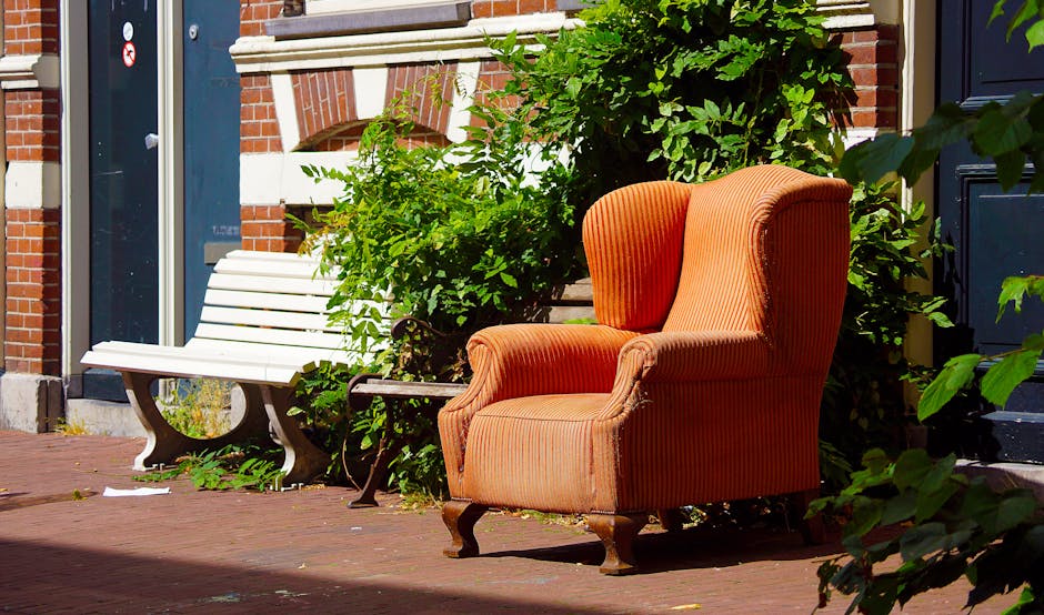 Outside a residential property in Mayfair, an orange upholstered armchair with carved wooden legs is positioned on the brick pavement beside a white wooden bench. Nearby, green foliage and shrubs partially obscure the view of the building’s brick and white stone exterior. The scene is illuminated by natural daylight, with shadows cast across the pavement. In the background, the building features a dark door and a window, suggesting the armchair and bench are situated on a front doorstep or alleyway. This setting reflects home relocation and furniture transport activities typical in house removals, with the furniture prepared for moving or for a new home. Man and Van Mayfair’s professional removals service might utilise such furniture during packing, loading, or delivery processes inside or outside properties, ensuring safe handling during furniture transport and home moving procedures.