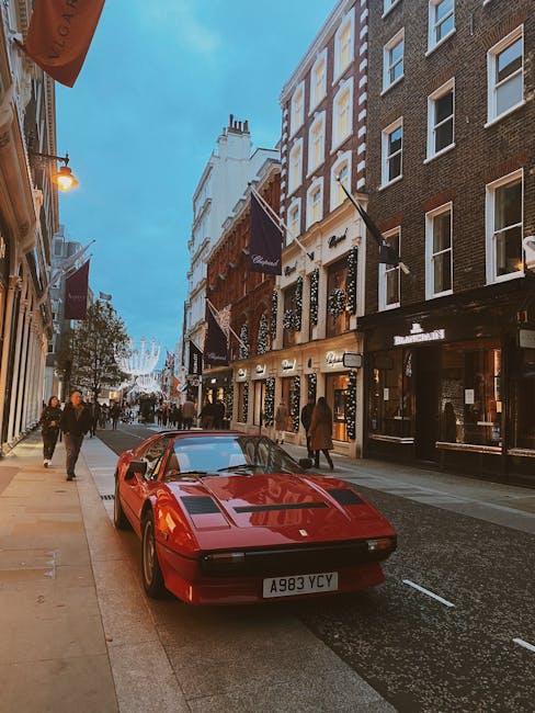 A red classic sports car, parked on a city street pavement near the curb, with a European-style number plate reading A983 YCY. Surrounding the vehicle are pedestrians walking along the sidewalk, some of whom are casual shoppers or residents. The street is lined with multi-story buildings featuring large windows, with signage and flags indicating shops and businesses, one of which is a luxury retail or hospitality venue. The buildings display a mix of modern and traditional architectural styles, with some having decorative awnings and ornate facades. Overhead, string lights are visible, suggesting a lively, urban environment, possibly during early evening or dusk. The scene reflects a busy city area suitable for home relocation and moving services like those provided by Man and Van Mayfair, with clear visibility of the street's layout and the surroundings involved in furniture transport or packing and moving logistics.
