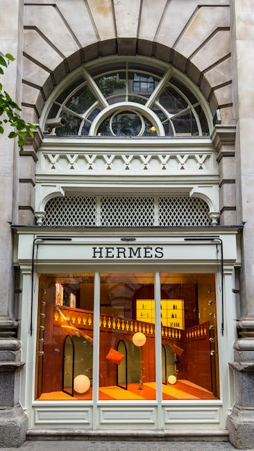 The image depicts the exterior of a building with an ornate stone facade and a large, arched window at the top. Below the arched window is a storefront with a white frame and a glass display area, showcasing high-end luxury items inside a Hermes boutique. The display window is illuminated with warm lighting, highlighting handbags and other accessories arranged on shelves. The storefront sign reads 'HERMES' in capital letters. The building's architecture features decorative molding and classical design elements, with a small balcony or decorative ledge above the shop entrance. Although the focus is on the exterior architecture of a retail location, the image can be associated with property and commercial fittings relevant to moving or refurbishment projects, such as packing and transportation of luxury goods during a home relocation or commercial move. This visually accurate description supports accessibility needs for individuals with visual impairments and aligns with the theme of high-value item transportation in professional removals.