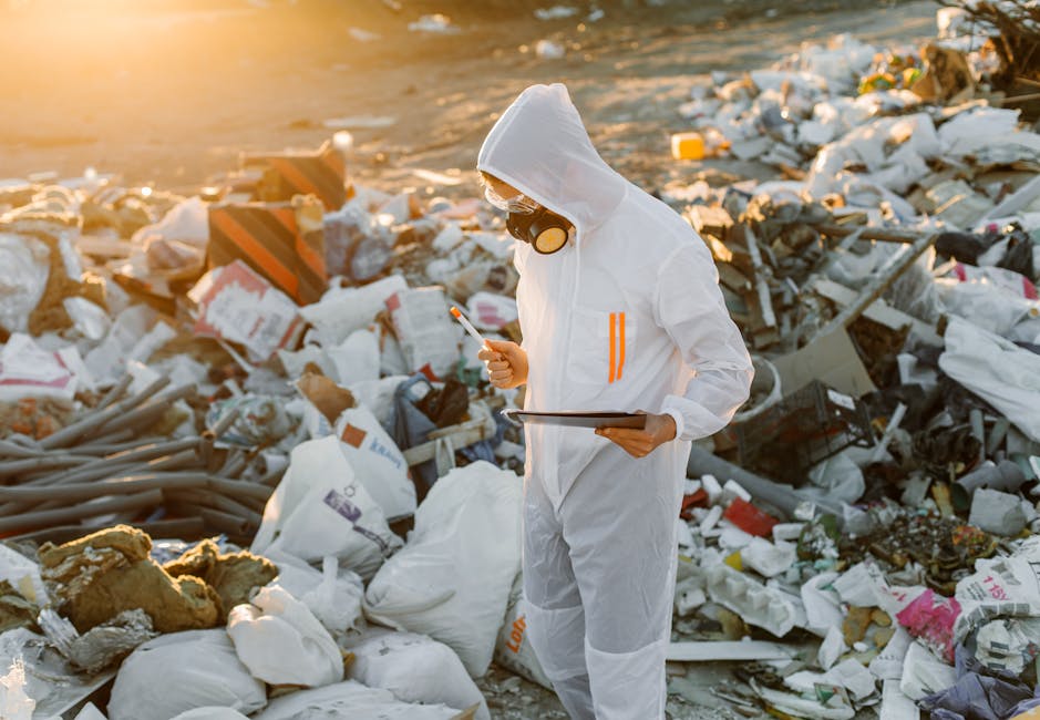 A person dressed in a white protective suit, gloves, and a face mask, standing amidst a large pile of mixed household waste, including plastic bags, cardboard boxes, foam insulation, and discarded household items, at an outdoor landfill site during sunset. The individual is holding a clipboard in one hand and a pen in the other, appearing to inspect or document the waste, with sunlight casting a warm glow over the scene. The environment suggests a focus on waste management or hazardous waste disposal, relevant to safe house and furniture removals. The setting reflects the importance of proper waste handling during home relocations to comply with hazardous waste rules, as discussed on the Man and Van Mayfair website.