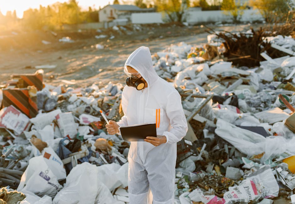 A person dressed in a white protective suit with orange reflective stripes, wearing a hood and safety goggles along with a mask, stands amidst a large pile of mixed waste including plastic bags, cardboard boxes, and debris on an outdoor landfill site during sunset. The individual is holding a clipboard in one hand and a pen in the other, inspecting or taking notes related to waste disposal. The background shows scattered rubbish extending towards a distant industrial or residential area, with warm sunlight illuminating the scene. This setting highlights the importance of proper waste management and hazardous waste disposal in the context of home relocation and moving services, with the image associated with the services of Man and Van Mayfair, who provide removals and transport solutions, including handling waste and packing materials during moves.