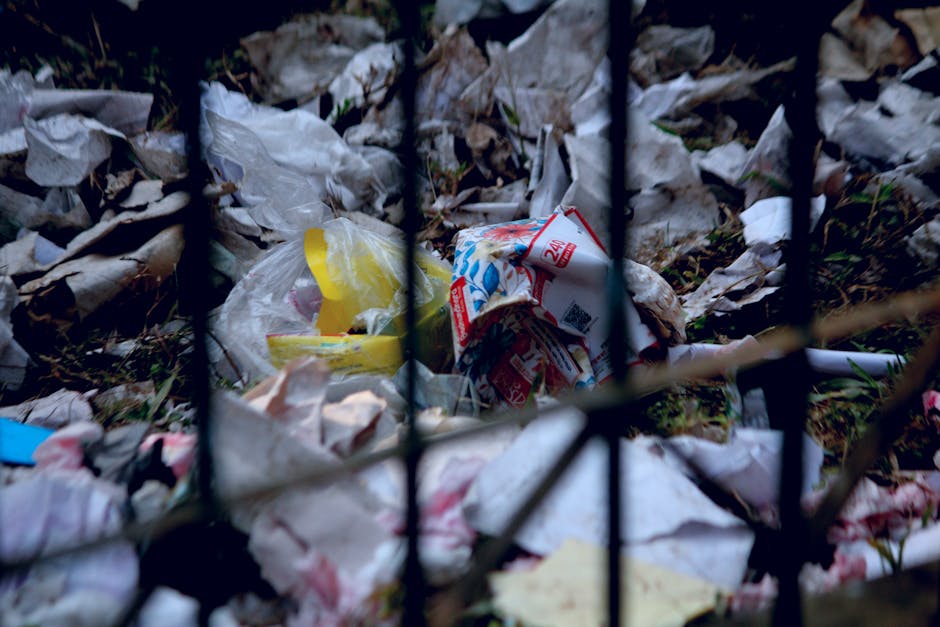 A person dressed in a white protective suit, gloves, and a face mask, standing amidst a large pile of mixed household waste, including plastic bags, cardboard boxes, foam insulation, and discarded household items, at an outdoor landfill site during sunset. The individual is holding a clipboard in one hand and a pen in the other, appearing to inspect or document the waste, with sunlight casting a warm glow over the scene. The environment suggests a focus on waste management or hazardous waste disposal, relevant to safe house and furniture removals. The setting reflects the importance of proper waste handling during home relocations to comply with hazardous waste rules, as discussed on the Man and Van Mayfair website.