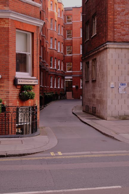 The image depicts a narrow residential street at the entrance of a red brick apartment building with multiple windows, situated at 36 Buckingham Gate. A small black wrought iron fence surrounds a potted flowering plant near the building's corner. The street curves slightly and leads to a set of stairs ascending between the buildings. Visible in the foreground are two yellow lines on the road, indicating parking restrictions. The scene appears to be part of a home relocation or furniture transport operation, possibly involving Man and Van Mayfair, who provide removals services. The natural lighting suggests daytime, with clear urban surroundings and the facade of the building providing a backdrop for moving activities such as loading or unloading household items within the vicinity of the property.