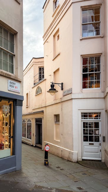 A narrow urban street scene showing the exterior of a multi-story sandstone building with multiple large, white-framed sash windows, some with visible interior furnishings through the glass. The building has a white-painted entrance door on the ground level and a traditional black wall-mounted lantern above. To the left, there is a storefront window displaying jewelry or accessories, with a sign reading 'Avery Howe.' The street is paved with grey cobblestones and features a bollard with a reflective orange strip, as well as a small round traffic sign. The sky is partially cloudy, and the scene suggests a quiet area suitable for house removals or furniture transport, as carried out by Man and Van Mayfair, specialists in removals involving tight spaces and narrow access points as indicated in the page about Mount Street Flats.