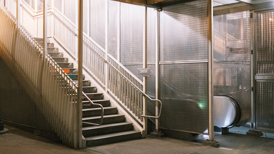 Interior view of a multi-storey building staircase with white metal railings leading up to an exit, positioned adjacent to a glass and metal enclosure housing an escalator. The staircase has black non-slip treads and is illuminated by natural light filtering through large windows or translucent panels on the upper level. On the landing, there is a small sign attached to the railing, and the area appears spacious with clean flooring. To the right side, the escalator's side panels and moving steps are partially visible, indicating potential movement of furniture or boxes during home relocation or packing and moving services. The environment suggests a typical urban setting used for building access, and this setup may present challenges for larger or bulky furniture transport, which is relevant to house removals specialists like Man and Van Mayfair, who often need to navigate such staircase and lift access issues during furniture transport or home removals.