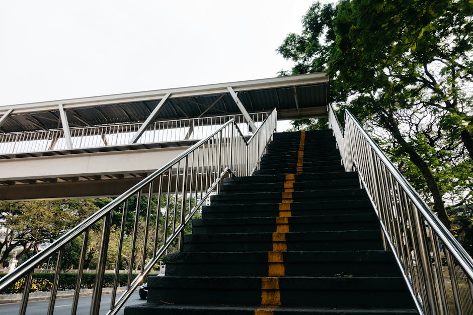 A set of outdoor metal stairs with black rubber treads and yellow safety markings running down the center, leading up to a bridge or elevated walkway beneath a cloudy sky. The stairs are flanked by silver metal handrails on both sides, and the surrounding environment includes green trees and foliage, indicating a park or landscaped area. The scene appears to be part of an urban public space or pedestrian crossing, potentially with challenges for home relocation or furniture transport due to stairs and limited access. In the context of house removals, Man and Van Mayfair may encounter these stairs during a moving process that involves navigating staircases and elevated walkways, requiring careful planning for loading and unloading of packing supplies, furniture, or boxes. The lighting suggests daytime, with natural light illuminating the scene, supporting the logistics involved in packing and moving across varied environments.