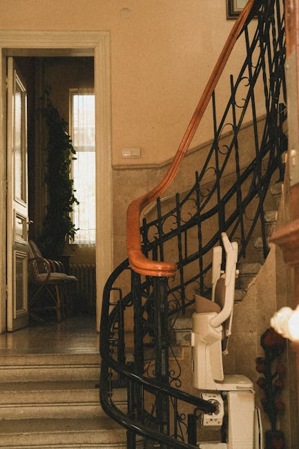 Interior view of a multi-storey building staircase with white metal railings leading up to an exit, positioned adjacent to a glass and metal enclosure housing an escalator. The staircase has black non-slip treads and is illuminated by natural light filtering through large windows or translucent panels on the upper level. On the landing, there is a small sign attached to the railing, and the area appears spacious with clean flooring. To the right side, the escalator's side panels and moving steps are partially visible, indicating potential movement of furniture or boxes during home relocation or packing and moving services. The environment suggests a typical urban setting used for building access, and this setup may present challenges for larger or bulky furniture transport, which is relevant to house removals specialists like Man and Van Mayfair, who often need to navigate such staircase and lift access issues during furniture transport or home removals.
