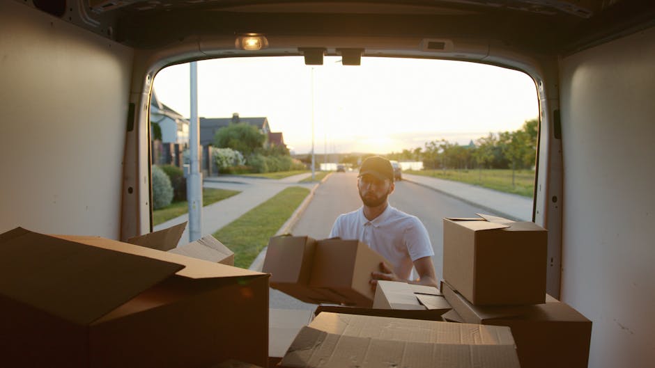 A man in a white polo shirt and sunglasses loading cardboard boxes into the back of a white van parked on a residential street during sunset. The open rear doors reveal several packed cardboard boxes of various sizes, some with flaps open, ready for a house removal or relocation process. The street features a paved sidewalk, green lawns, and houses with trees and bushes in the background, under warm golden lighting. The image captures the loading process during a home relocation, with Man and Van Mayfair providing moving services, emphasizing packing and furniture transport as part of their removal offerings.