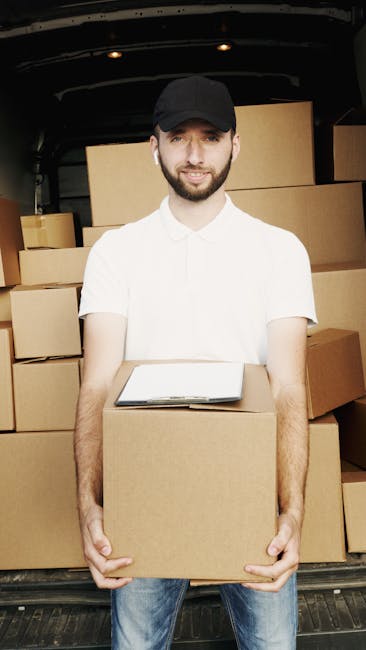 A man with a beard and wearing a black cap and white polo shirt is standing outside next to an open moving van, holding a cardboard box. The van's interior features neatly packed cardboard boxes stacked on the right side, some wrapped in plastic, ready for transport as part of a home relocation process. The scene takes place outdoors during daylight, with greenery and a body of water visible in the background, indicating a suburban or rural setting. The man appears prepared for the loading process, which involves lifting, carrying, and loading items into the van, reflecting professional furniture transport and packing activities associated with house removals. The setting highlights the logistics involved in last-minute booking for house moves in Mayfair, as managed by Man and Van Mayfair.