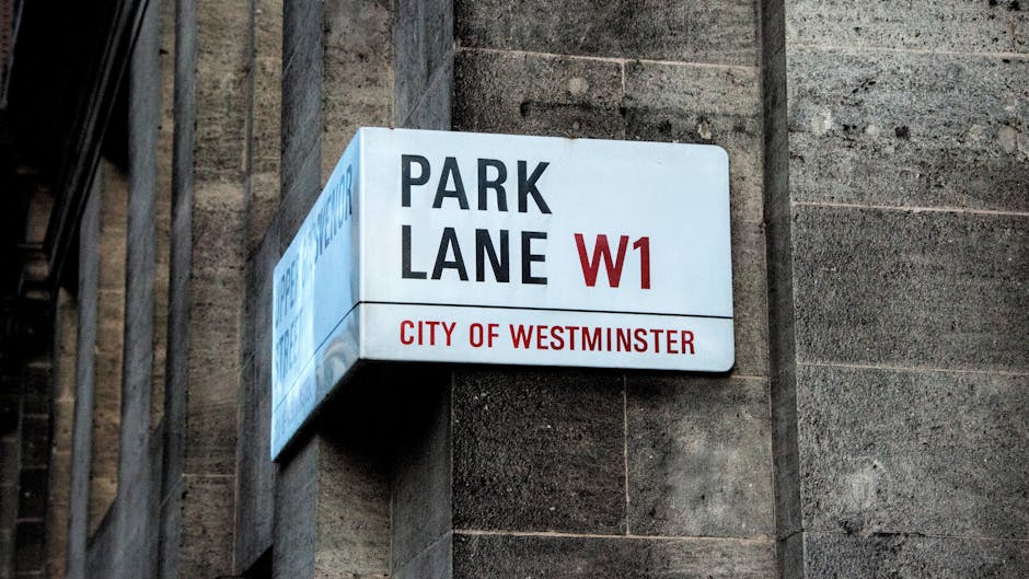 A rectangular street sign mounted on a dark brick wall in Westminster, London, displaying the text 'PARK LANE W1' in large black and red letters, with 'CITY OF WESTMINSTER' written beneath in red. The sign is illuminated and positioned at a height, with visible metal brackets supporting it. The surrounding environment features aged brickwork and metal railings, indicating an urban setting. This image relates to house removals and moving logistics within Mayfair, highlighting the importance of permits for parking and loading during furniture transport and home relocation processes facilitated by companies like Man and Van Mayfair.