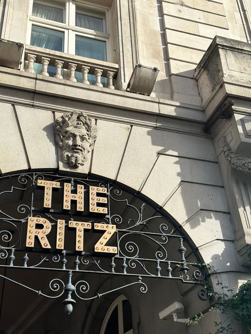 Close-up view of the exterior of a historic building with a decorative stone face sculpture above a black iron gate featuring intricate scrollwork and a bright, illuminated sign spelling 'THE RITZ' with large, round yellow bulbs. The building is made of light-colored stone with architectural details such as a window with a decorative balustrade, a small protruding ledge, and stone masonry accents on the façade. The scene is lit by natural daylight, highlighting the textured stone surfaces and casting shadows. This setting may be part of an urban area where professional removals, such as packing, furniture transport, or home relocation services provided by companies like Man and Van Mayfair, are required for moving or delivery logistics within historic city buildings.